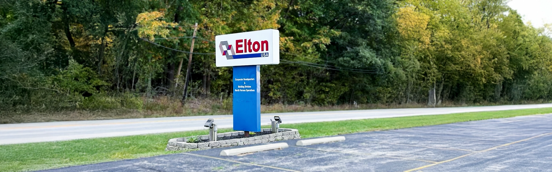 A tall, modern outdoor sign for "Elton USA" featuring a red "E" logo, positioned on a grassy median next to an empty parking lot and a road, with a dense line of trees in the background.